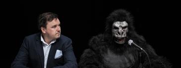 Man in a suit sitting beside a fake gorilla. 
